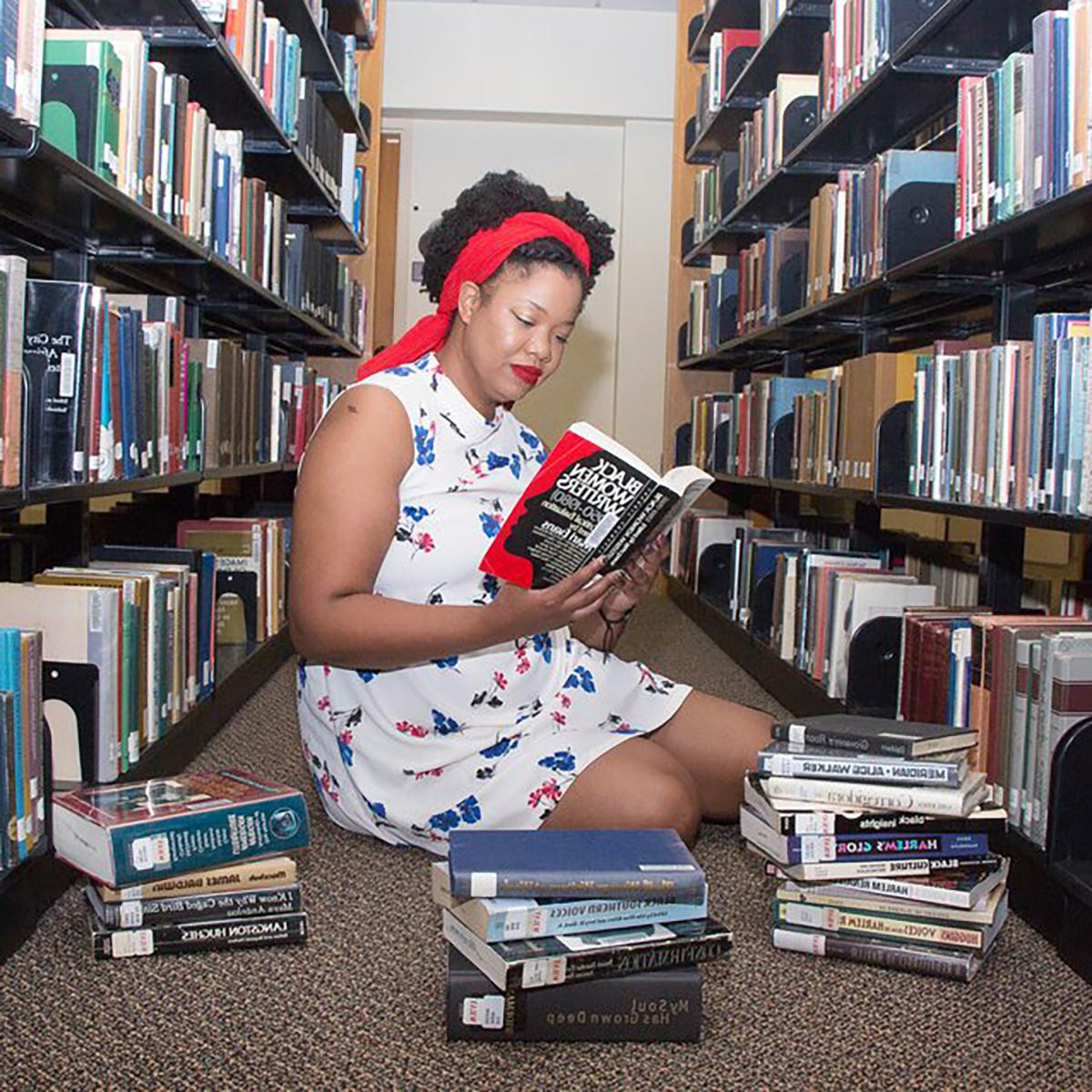 凯特琳·亨特摄, 年轻的黑人妇女, seated between two library bookshelves with a pile of books in front of her. She is reading a book titled "Black Women Writers, 1950-1980"