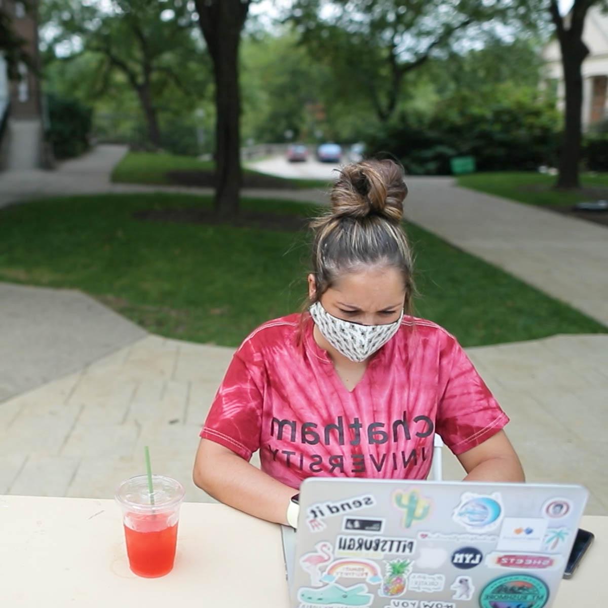 Photo of a young female student in a tie dye Chatham University shirt, wearing a mask 和 seated at a table in the quad on 足球波胆平台